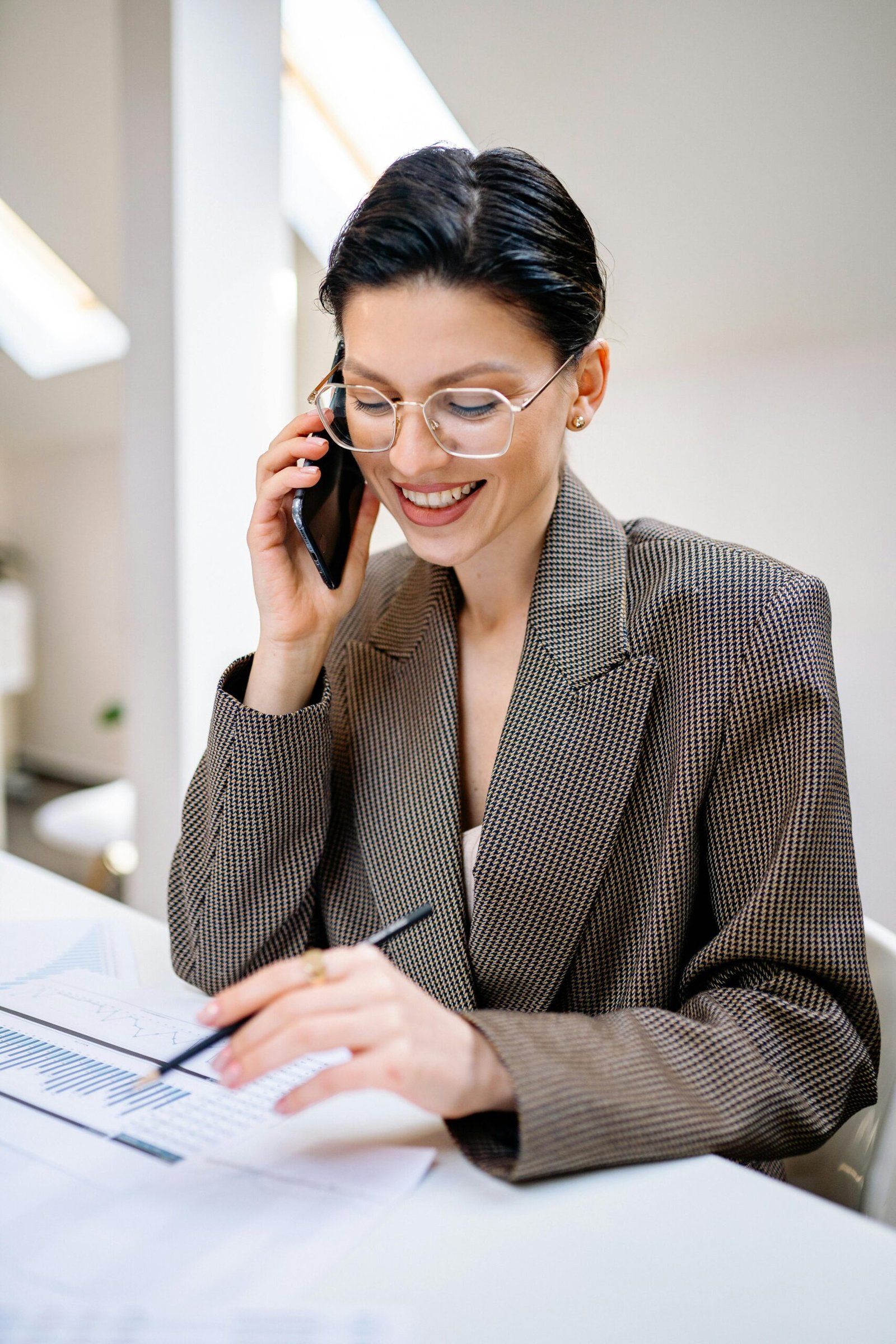 pexels-photo-7693223-7693223 Confident businesswoman wearing glasses, talking on phone while reviewing documents in modern office setting.